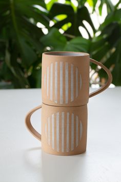 two coffee mugs sitting next to each other on top of a white table with green plants in the background