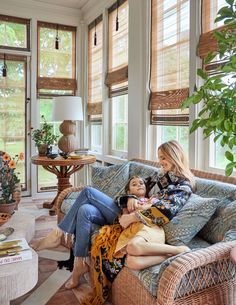 two women sitting on a couch in a living room with large windows and wicker furniture