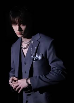 a man in a suit and chain necklaces standing against a black background with his hand on his chest