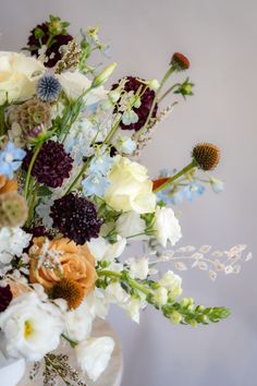 deep purple, orange, white, brown and blue fall floral arrangement in a white container in front of a white wall Fall Floral Arrangements With Blue, White And Brown Flower Arrangements, Scabiosa Arrangement, October Flower Arrangements, Fall Wedding Flowers September, October Flowers In Season, Delphinium Wedding Bouquet, Purple Scabiosa, Autumn Flower Arrangements