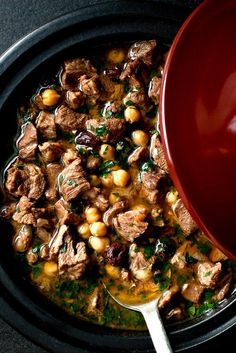 a red bowl filled with meat and chickpeas next to a white spoon on a black surface