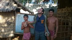 three men standing in front of a hut with a snake on his arm and another man holding an animal