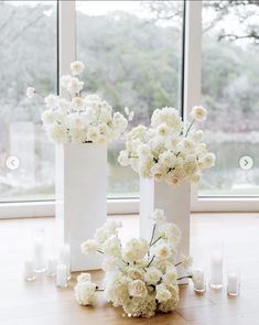 three white vases filled with flowers sitting on top of a table next to candles