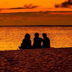 three people sitting on the beach at sunset