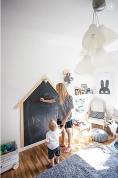 a woman and child writing on a blackboard in a room with white walls, hardwood floors and blue rugs