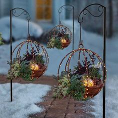 three metal ornaments with lights hanging from them on poles in the snow, surrounded by grass and plants