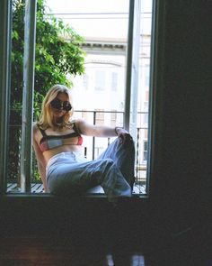 a woman sitting in front of a window with her legs crossed and wearing sunglasses on