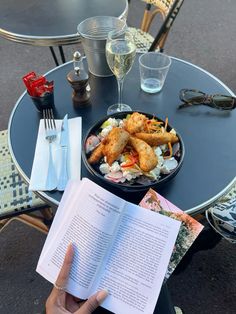 a person is reading a book at an outdoor table with food and glasses on it