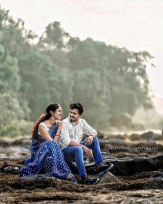 a man and woman sitting next to each other on rocks near the water with trees in the background