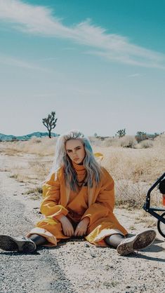 a woman sitting on the side of a dirt road next to a bike and trailer