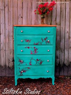 a blue dresser with flowers painted on the top and bottom, sitting in front of a wooden fence