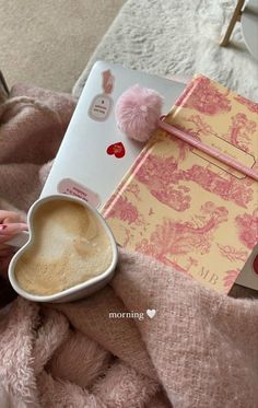 a cup of coffee sitting on top of a bed next to an open book and pink pom - pom