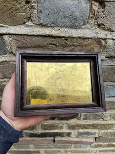 a hand holding up a small wooden frame with a painting on it in front of a brick wall