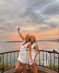 two beautiful young women standing next to each other near the ocean at sunset with their arms in the air