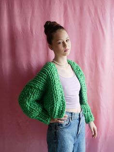 a woman standing in front of a pink wall wearing a green cardigan sweater and jeans