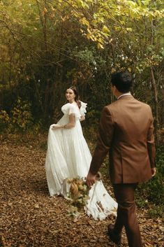 the bride and groom are walking through the woods in their wedding attire, holding hands