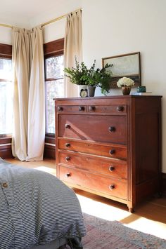 a bedroom scene with focus on the dresser and bed in the foreground, and sunlight coming through the window