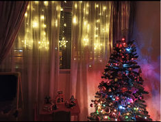 a christmas tree is lit up in front of a window with curtains and lights behind it