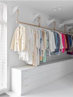 an organized closet with clothes hanging on the rack and white drawers below it, along with open shutters