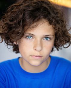 a young boy with curly hair and blue eyes looks at the camera while wearing a blue t - shirt