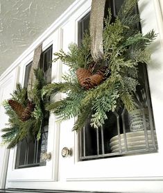 a wreath with pine cones and evergreen leaves hanging on the front door window sill