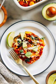 an enchilada on a white plate with a fork and avocado next to it