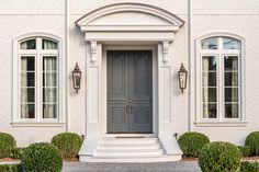 the front door of a white house with two large windows and trimmed bushes on either side