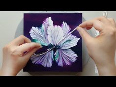 two hands are painting a flower on a purple and white square with watercolors