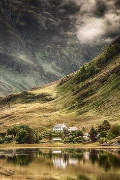 a house on the side of a mountain with water in front of it and clouds above