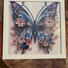 a butterfly made out of beads and flowers in a white frame on a wooden table