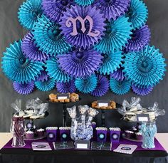 a purple and blue dessert table set up with paper fans on the wall behind it