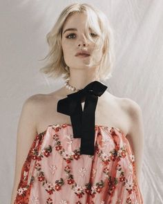 a woman with blonde hair wearing a pink floral dress and black bow neck tie, standing in front of a white backdrop