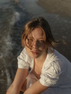 a woman in white shirt sitting on beach next to water and looking at the camera