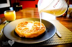 a waffle on a plate with syrup being poured over it and orange juice in the background