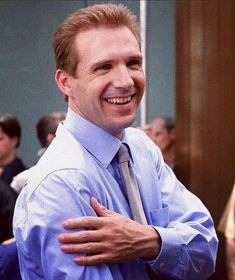 a man in a blue shirt and tie smiles while standing with his arms around his chest