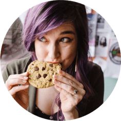 a woman with purple hair is eating a chocolate chip cookie in front of her face