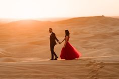 a man and woman in formal wear walking across sand dunes at sunset with the sun behind them