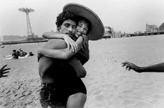 two people hug on the beach while others play in the water and sand behind them