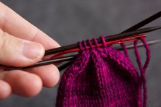 a hand holding two knitting needles over a purple piece of cloth with red yarn on it