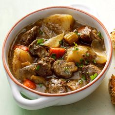 a white bowl filled with meat and vegetables next to some bread on a counter top