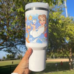 a person holding up a disney princess travel mug in front of some trees and grass