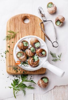 small snails in a white bowl on a wooden cutting board next to garlic and parsley