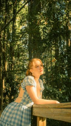 a woman in a blue and white dress leaning on a wooden fence with trees behind her