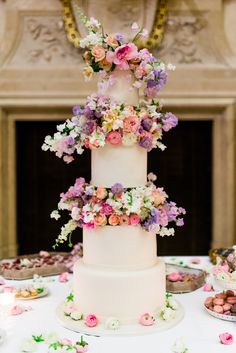 a three tiered wedding cake with colorful flowers on the top and bottom, sitting on a table in front of a fireplace