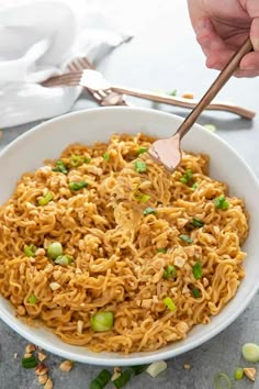 a person holding a wooden spoon over a bowl of noodles with peas and peanuts on the side