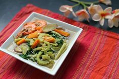 a white bowl filled with noodles and vegetables on top of a red table cloth next to flowers