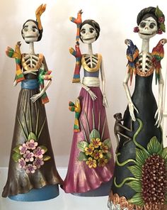 three ceramic figurines in the shape of skeletons with flowers and birds on their heads