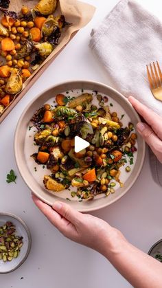 two hands holding a plate with roasted vegetables on it, next to other plates and silverware