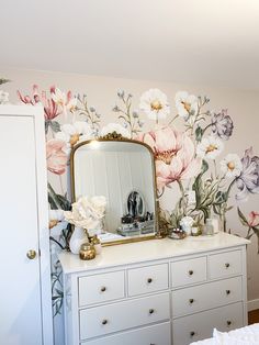 a white dresser topped with a mirror next to a wall covered in floral wallpaper