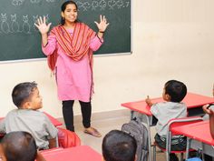 a woman standing in front of a classroom full of children sitting at desks and holding their hands up